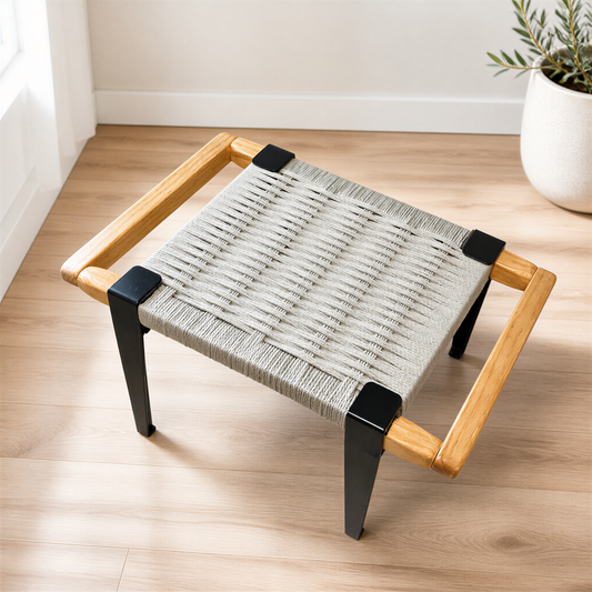 A woven ottoman with a wooden frame sits on a wooden floor, accompanied by a potted plant in the background.