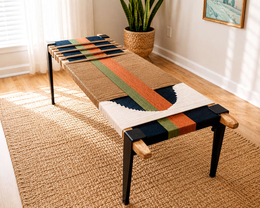 a woven bench with a colorful, abstract design on the seat and legs, placed on a woven rug in a room with a window and a plant.