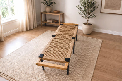a cozy living room with a woven bench, a rug, and a potted plant. The room has a warm and inviting atmosphere, with natural light coming in from a window.