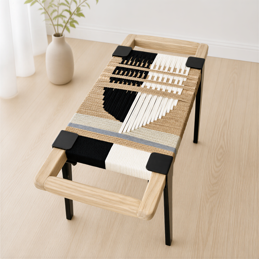 a modern, minimalist coffee table made of woven materials in black, white, and beige colors. The table has a wooden frame and a curved, wooden handle. It is placed on a wooden floor, and there is a vase with a plant in the background.