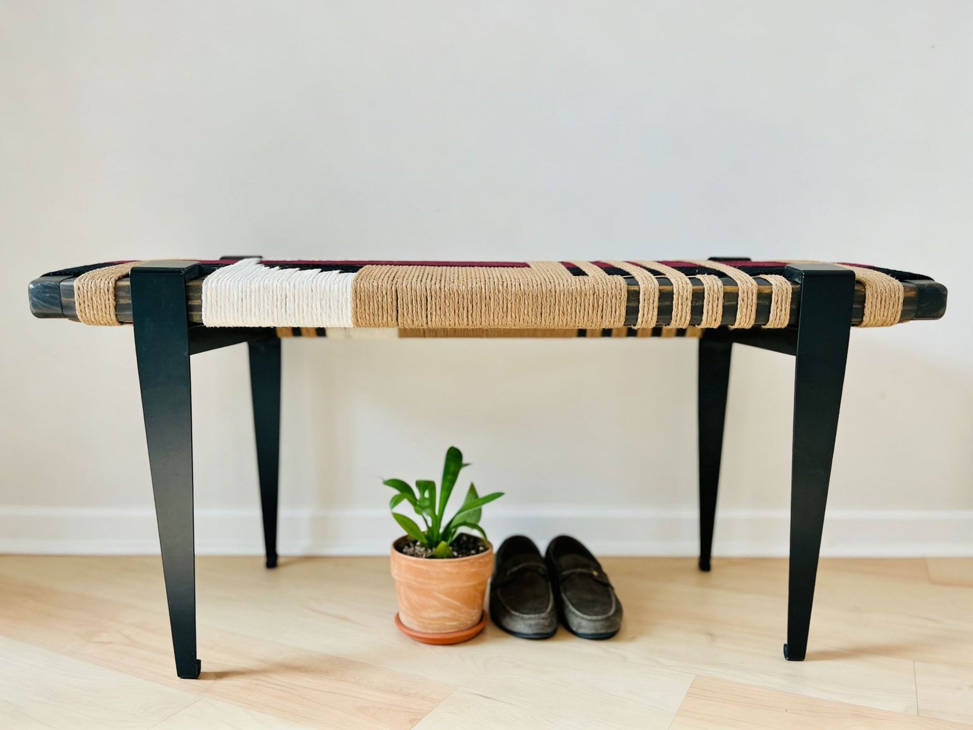 A modern bench with a woven seat and black metal legs, placed on a wooden floor next to a potted plant and a pair of shoes.