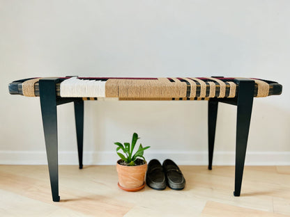A modern bench with a woven seat and black metal legs, placed on a wooden floor next to a potted plant and a pair of shoes.