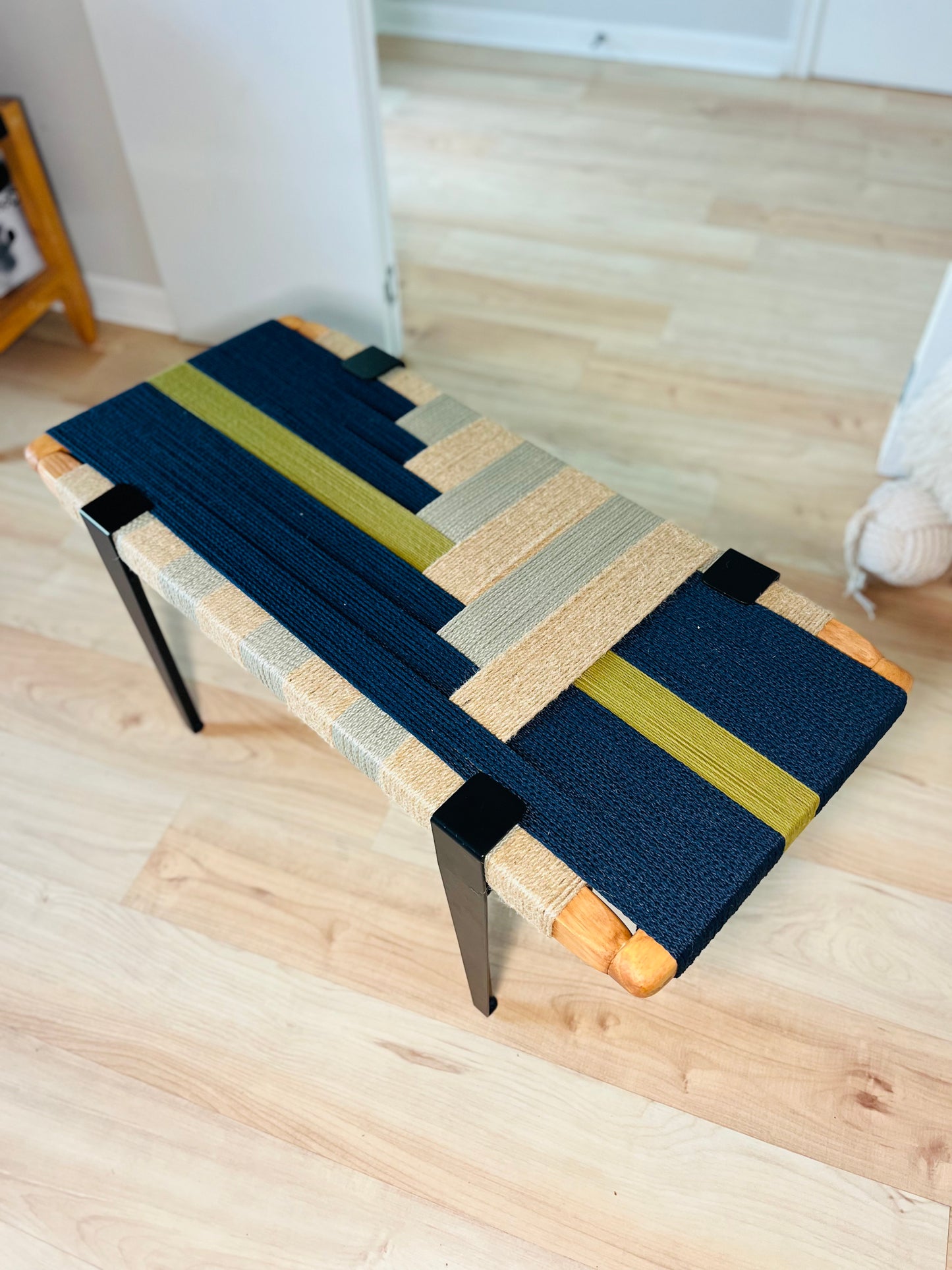 a wooden bench with a colorful woven seat and legs, placed on a wooden floor.