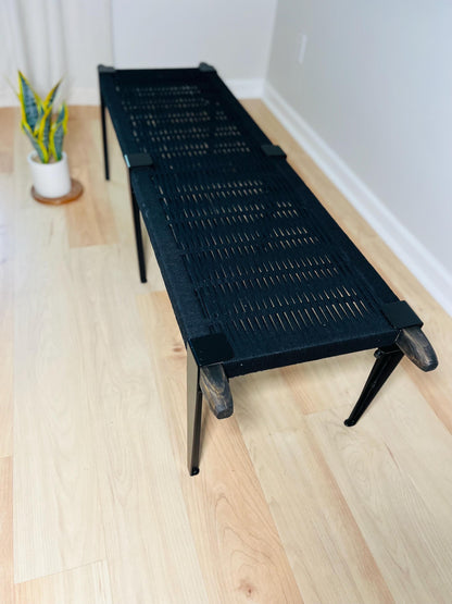 A black woven bench with a woven seat and legs sits on a wooden floor, accompanied by a potted plant in the background.