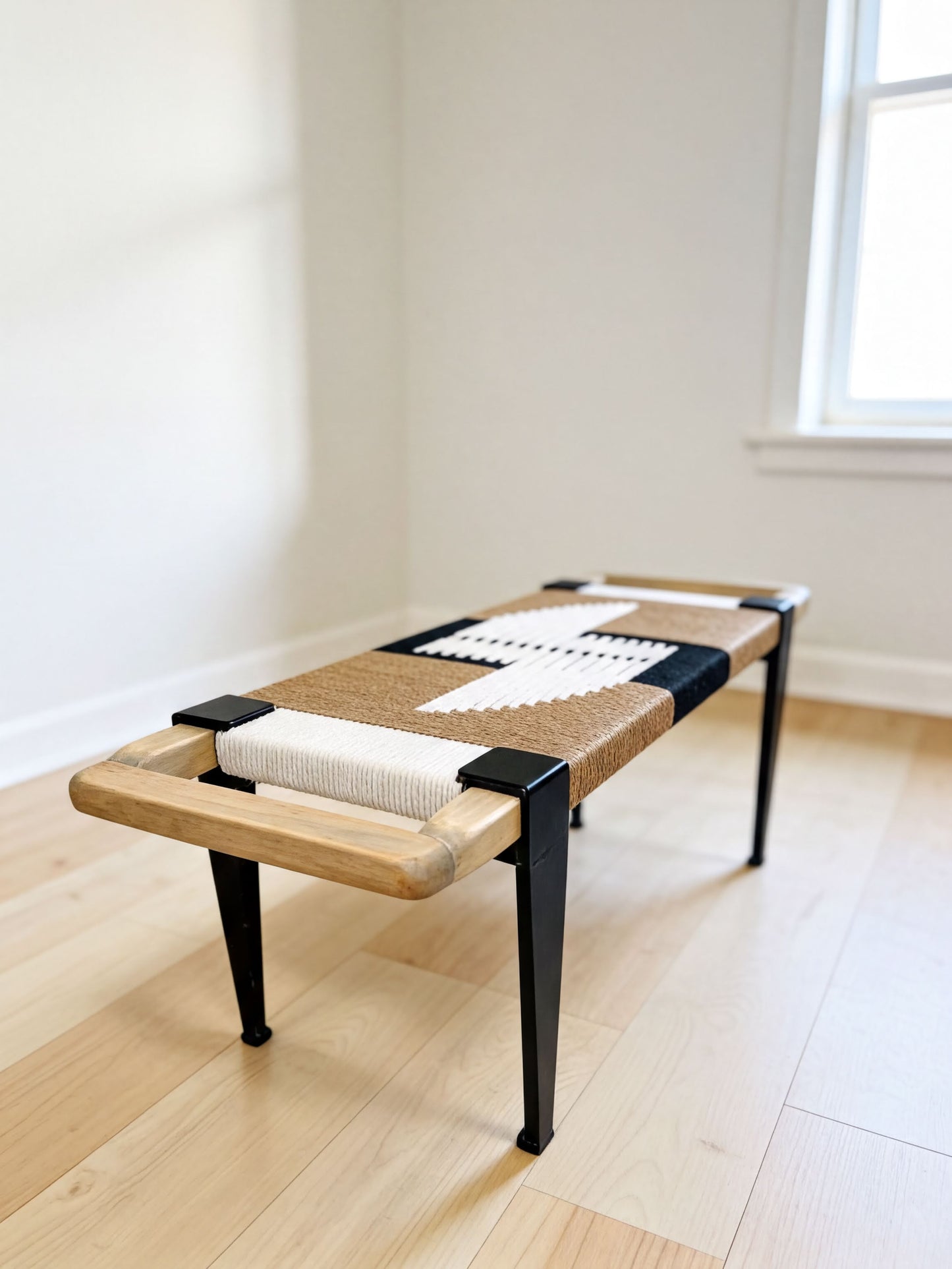 A wooden bench with a woven seat and black metal legs, placed on a wooden floor against a white wall.