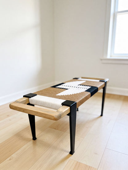 A wooden bench with a woven seat and black metal legs, placed on a wooden floor against a white wall.