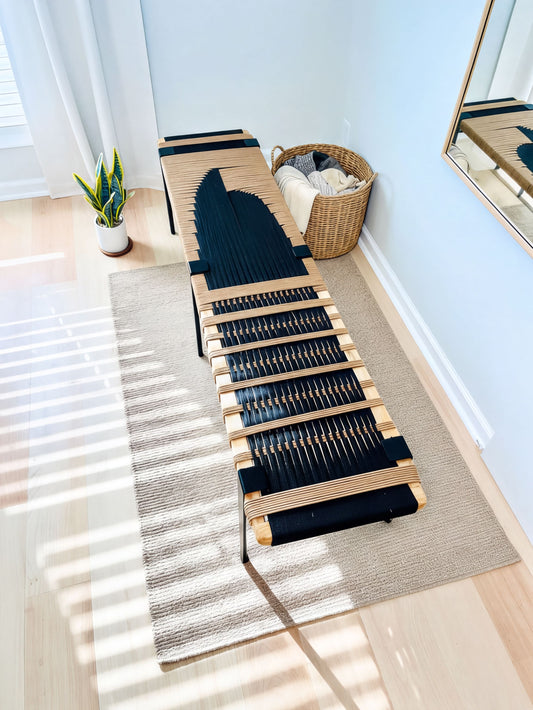 a modern black and brown bench with a woven seat, placed on a beige rug in a room with a window and a plant.