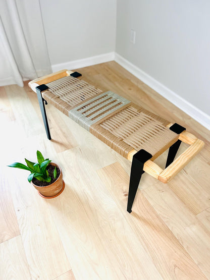 a woven bench with black metal legs, placed on a wooden floor next to a potted plant.