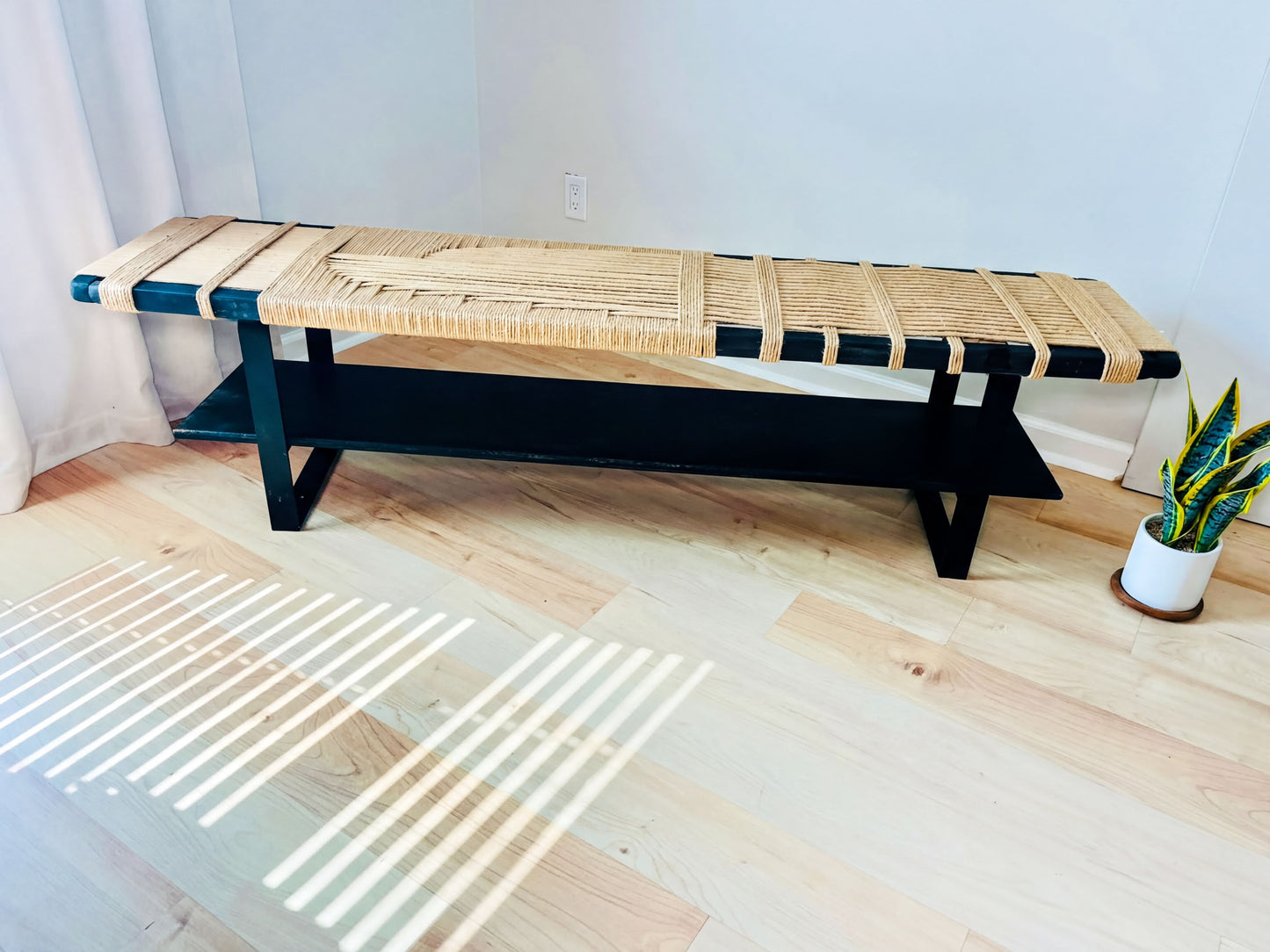 a black bench with a woven seat and a black shelf, placed on a wooden floor. There is a potted plant next to the bench, adding a touch of greenery to the scene.