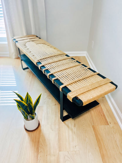 a modern bench with a woven seat and black metal frame, placed on a wooden floor next to a potted plant.