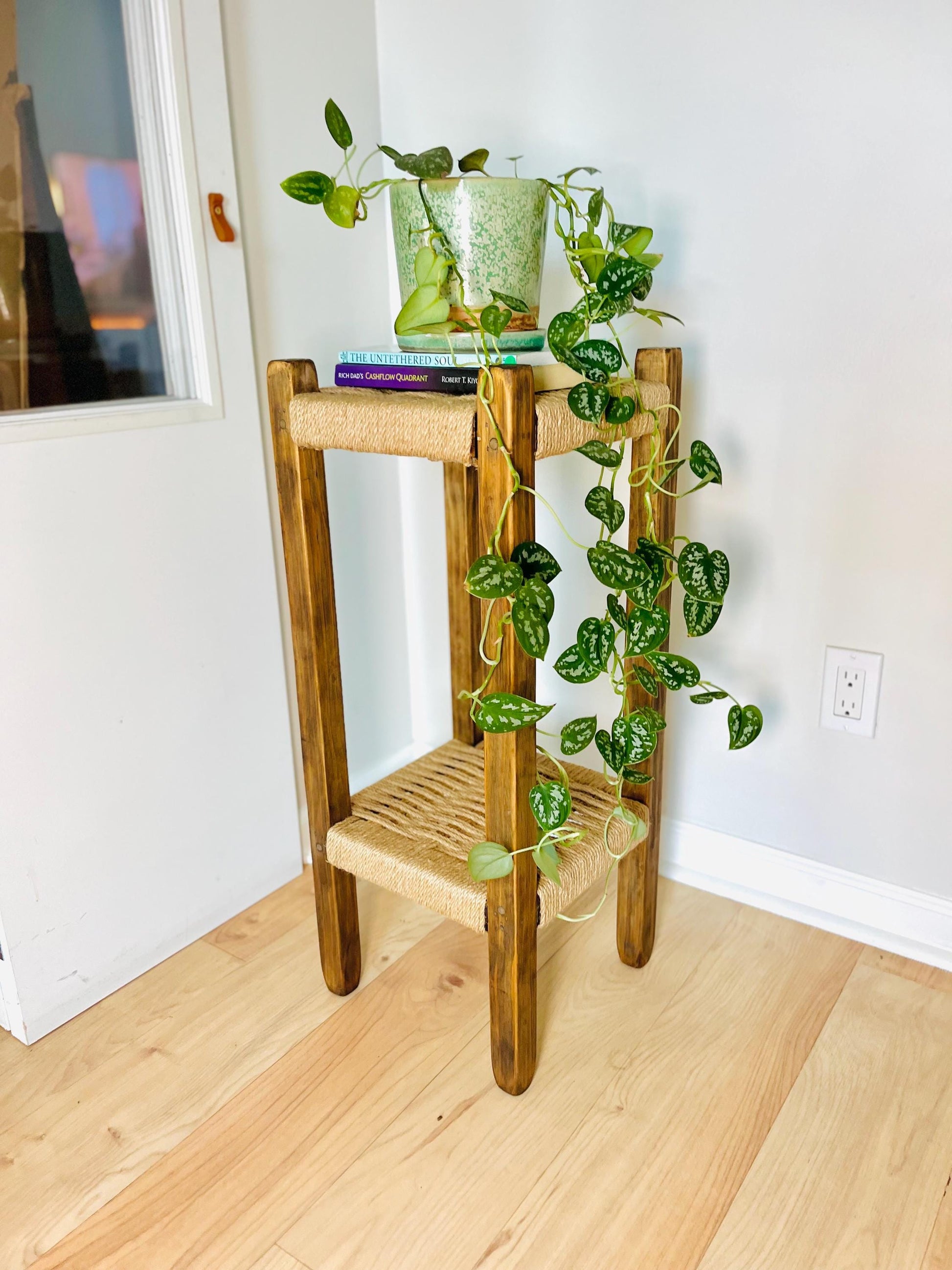 A wooden side table with a woven seat and a plant hanging from the top, placed on a wooden floor against a white wall.