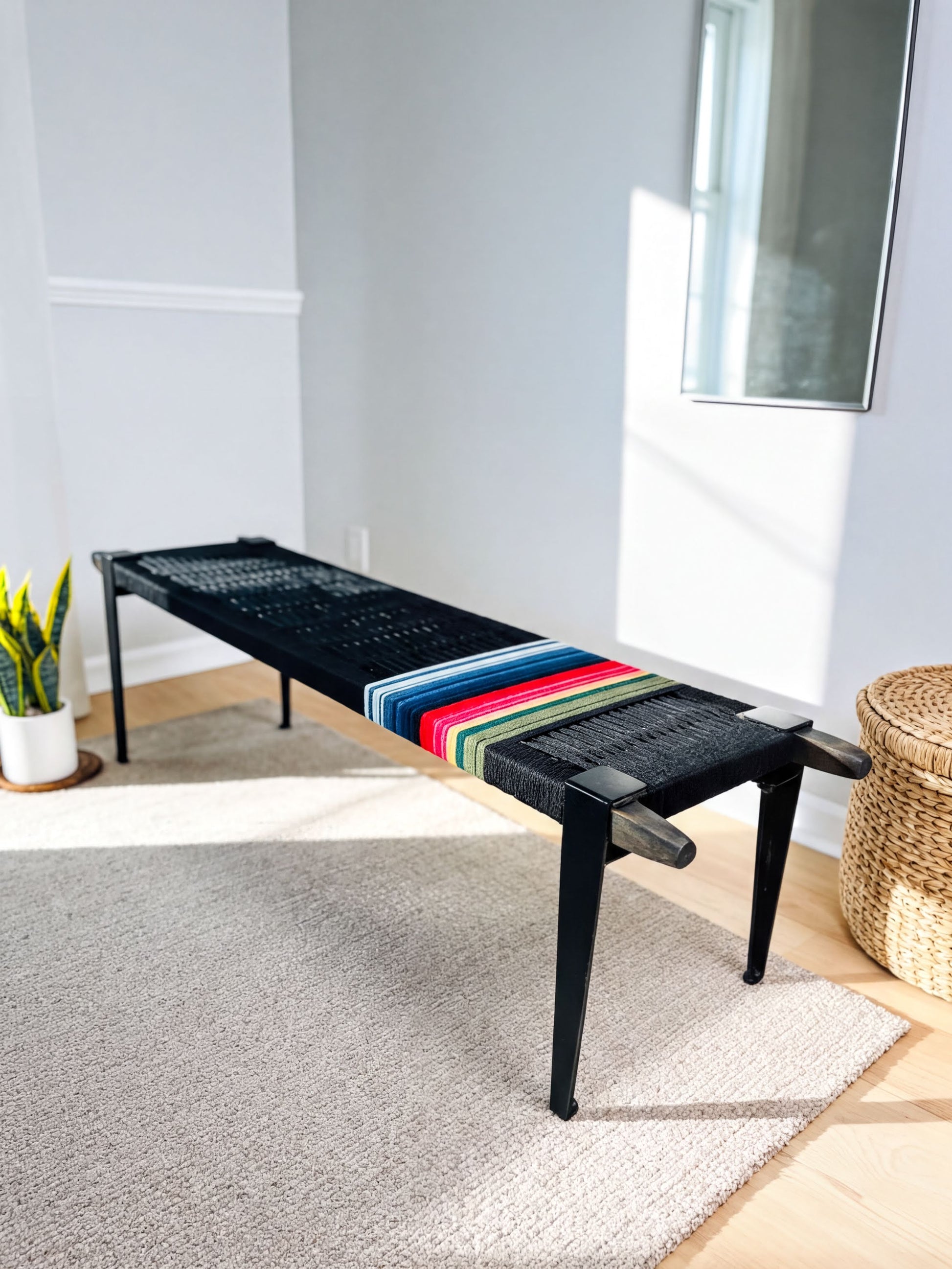 a black bench with a colorful striped seat, placed on a gray rug in a room with white walls and a window.