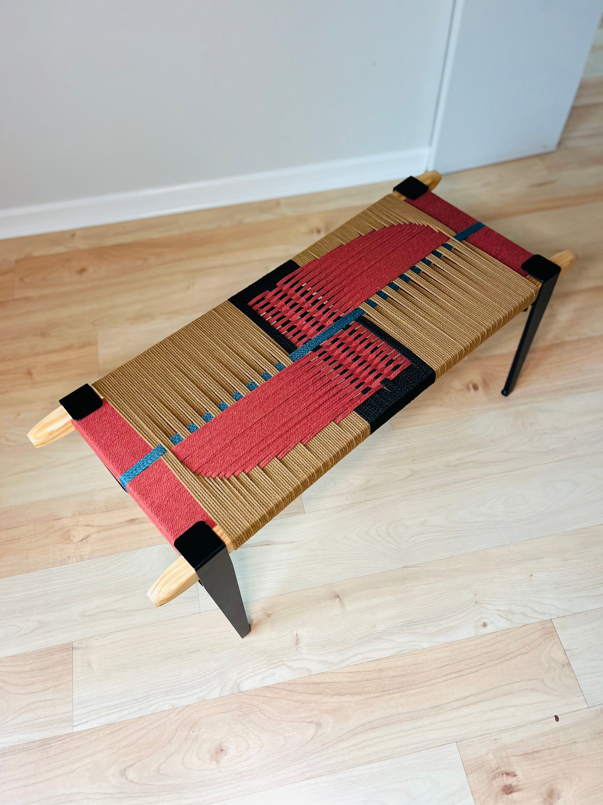 A woven bench with a red and black geometric pattern on the seat and legs, placed on a wooden floor against a white wall.