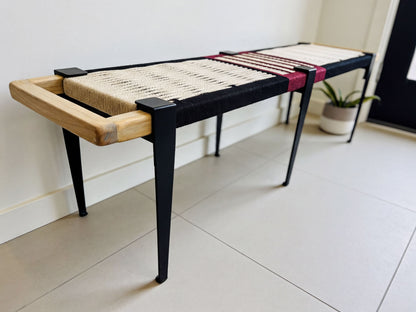 A modern bench with a black metal frame and a woven seat, placed on a tiled floor against a white wall.