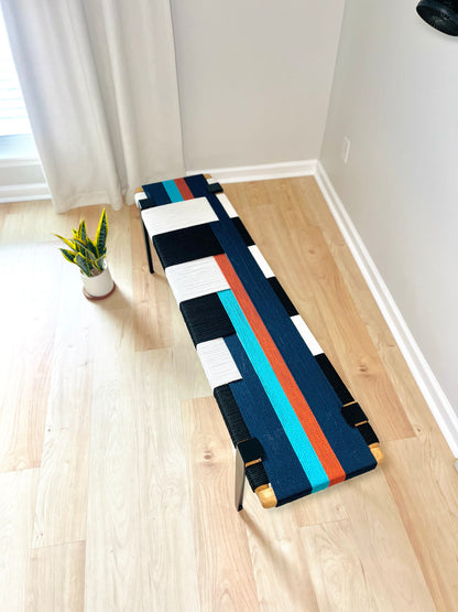 a colorful bench with a geometric pattern of blue, white, and orange stripes, placed on a wooden floor next to a window.