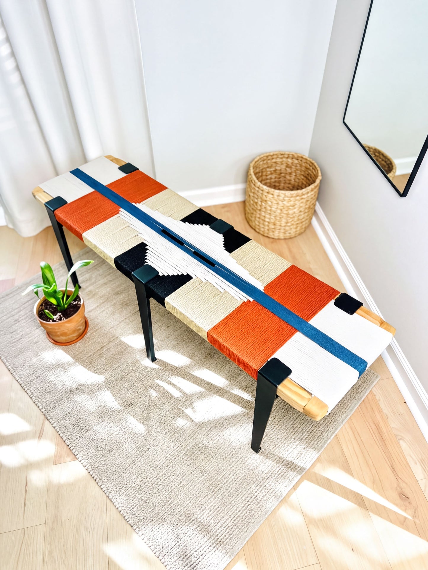 a colorful bench with a woven seat and legs, placed on a rug in a room with a mirror and a potted plant nearby.