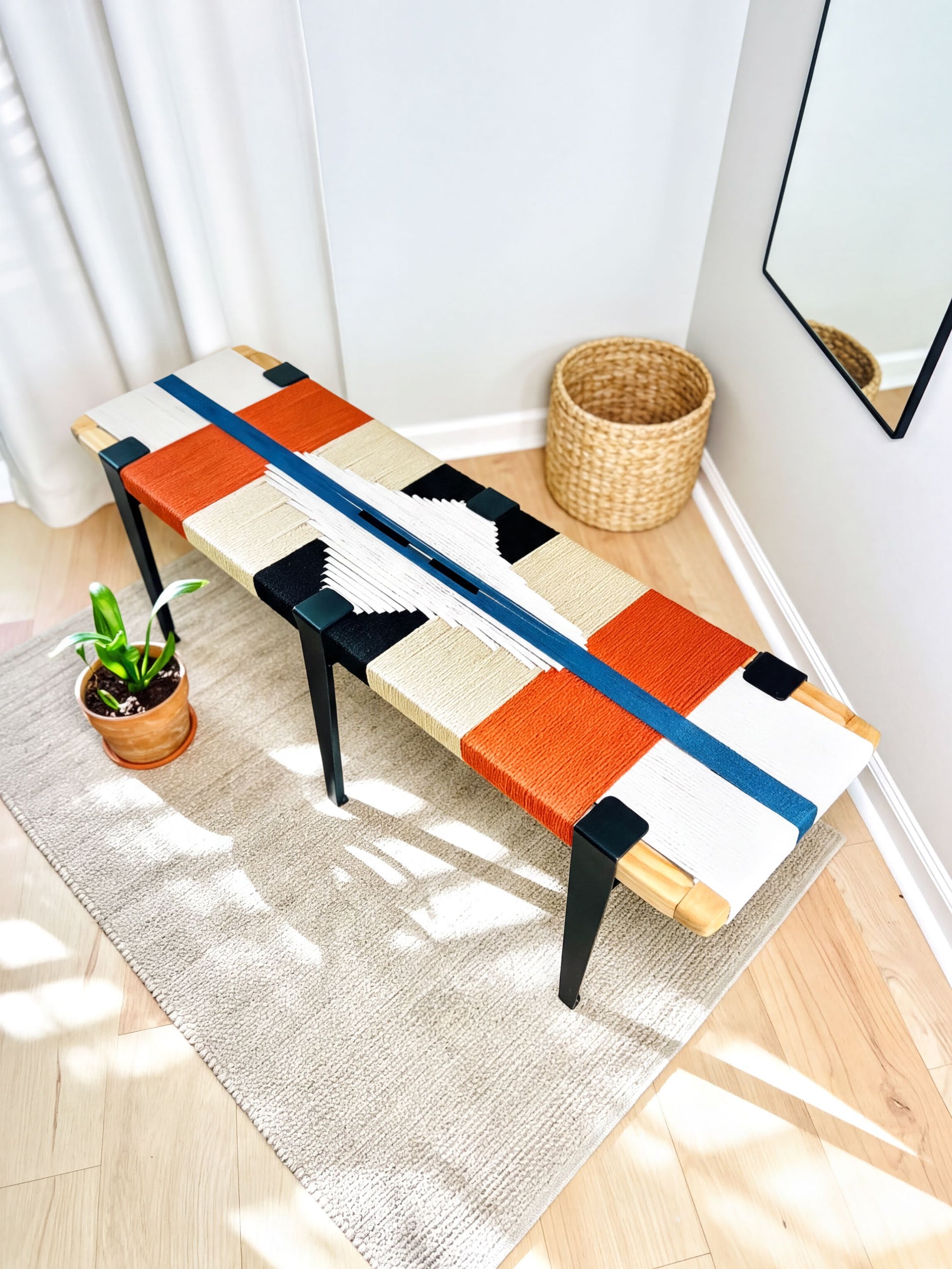 a colorful bench with a woven seat and legs, placed on a rug in a room with a mirror and a potted plant nearby.