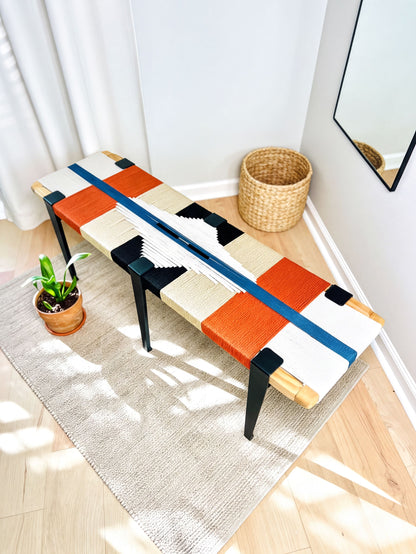 a colorful bench with a woven seat and legs, placed on a rug in a room with a mirror and a potted plant nearby.
