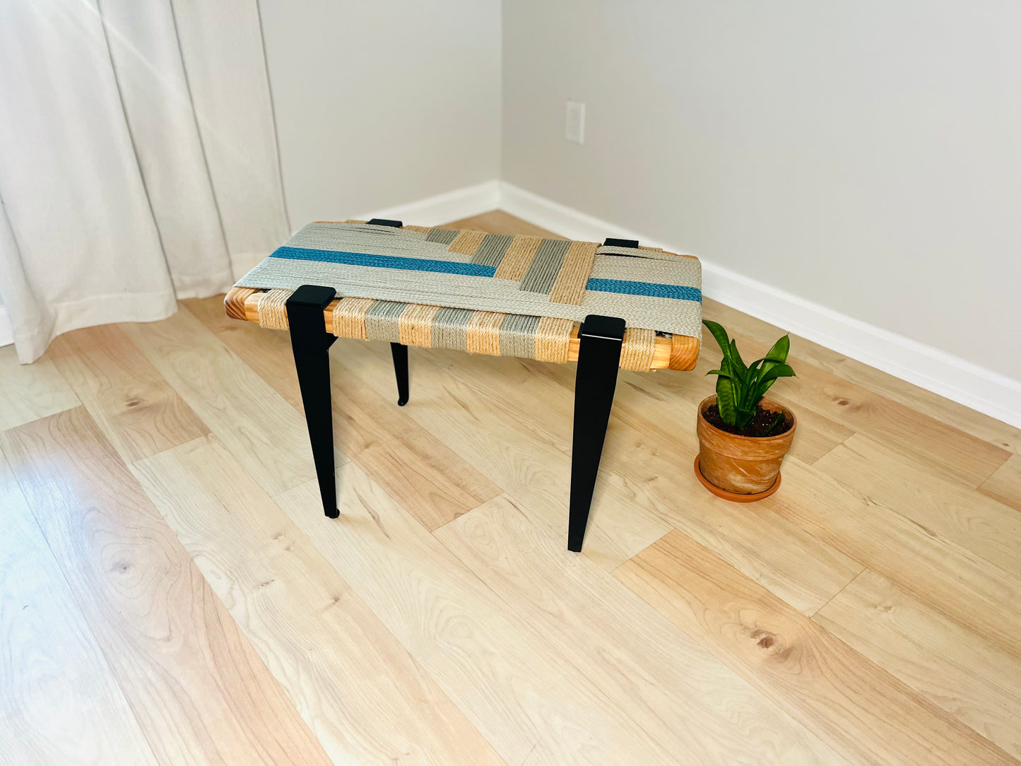 a wooden bench with a woven seat and black metal legs, placed on a wooden floor next to a potted plant.