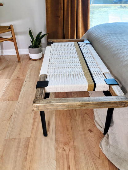 a bedroom with a wooden bench placed on a hardwood floor. The bench has a woven seat and a wooden frame, and it is positioned next to a bed. There is a chair and a potted plant in the room, adding to the cozy and inviting atmosphere.