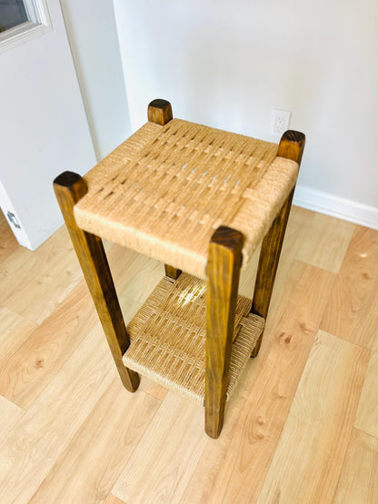 a wooden side table with a woven seat and two wooden legs, placed on a wooden floor against a white wall.