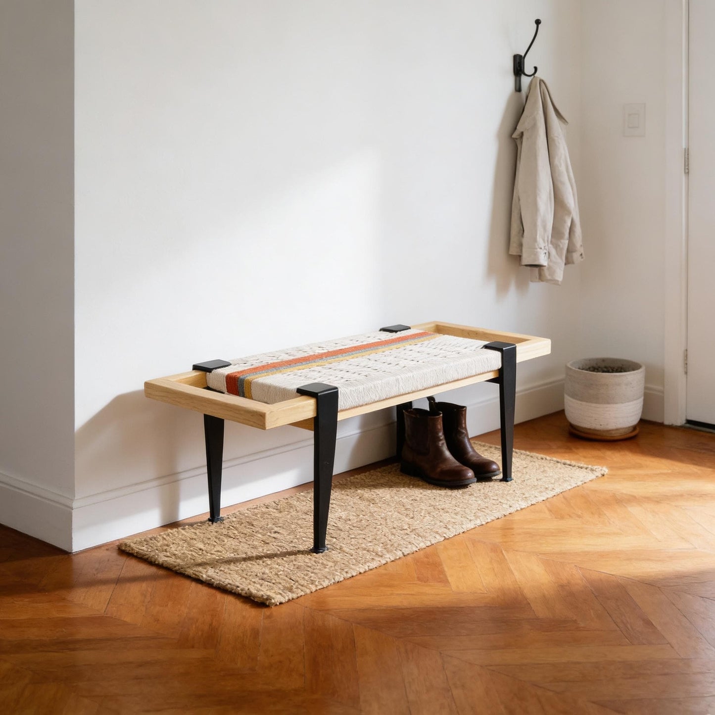 a wooden bench with black metal legs, placed on a rug in a hallway. A coat is hanging on a hook nearby, and a pair of brown boots is resting on the rug.