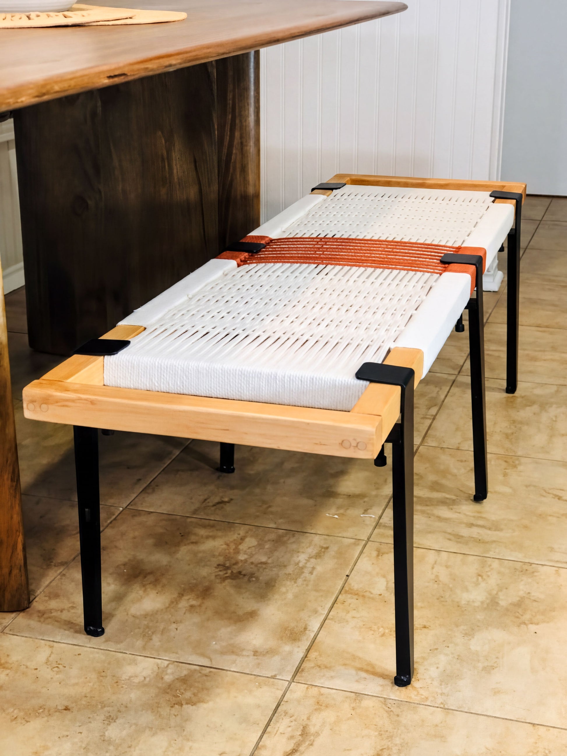 a wooden bench with a woven seat and black metal legs, placed on a tiled floor.