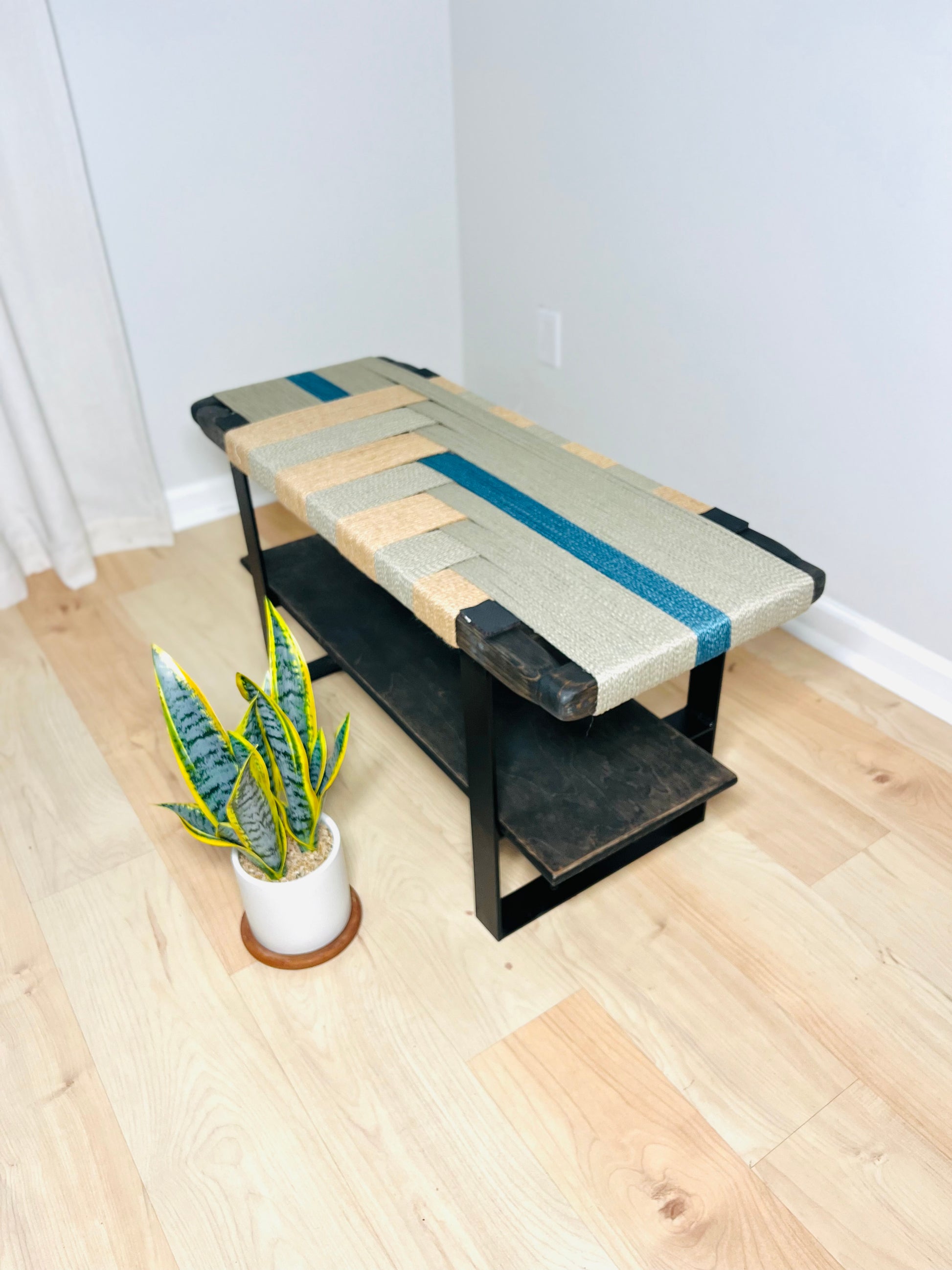 a wooden bench with a woven seat and a black metal frame, placed on a wooden floor next to a potted plant.