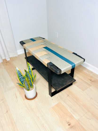 a wooden bench with a woven seat and a black metal frame, placed on a wooden floor next to a potted plant.
