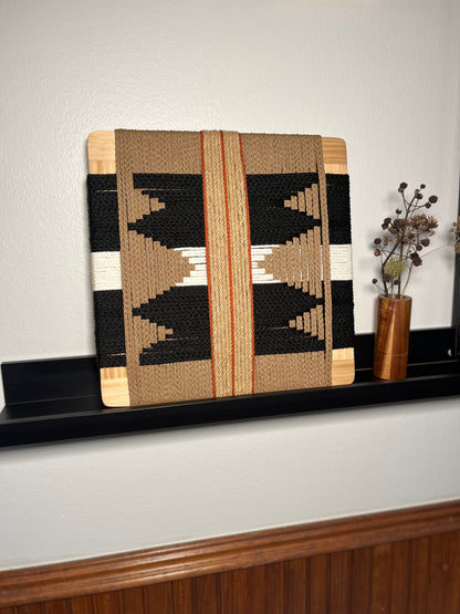 a wooden shelf displaying a woven piece of art, a vase with dried flowers, and a wooden board with a geometric pattern.