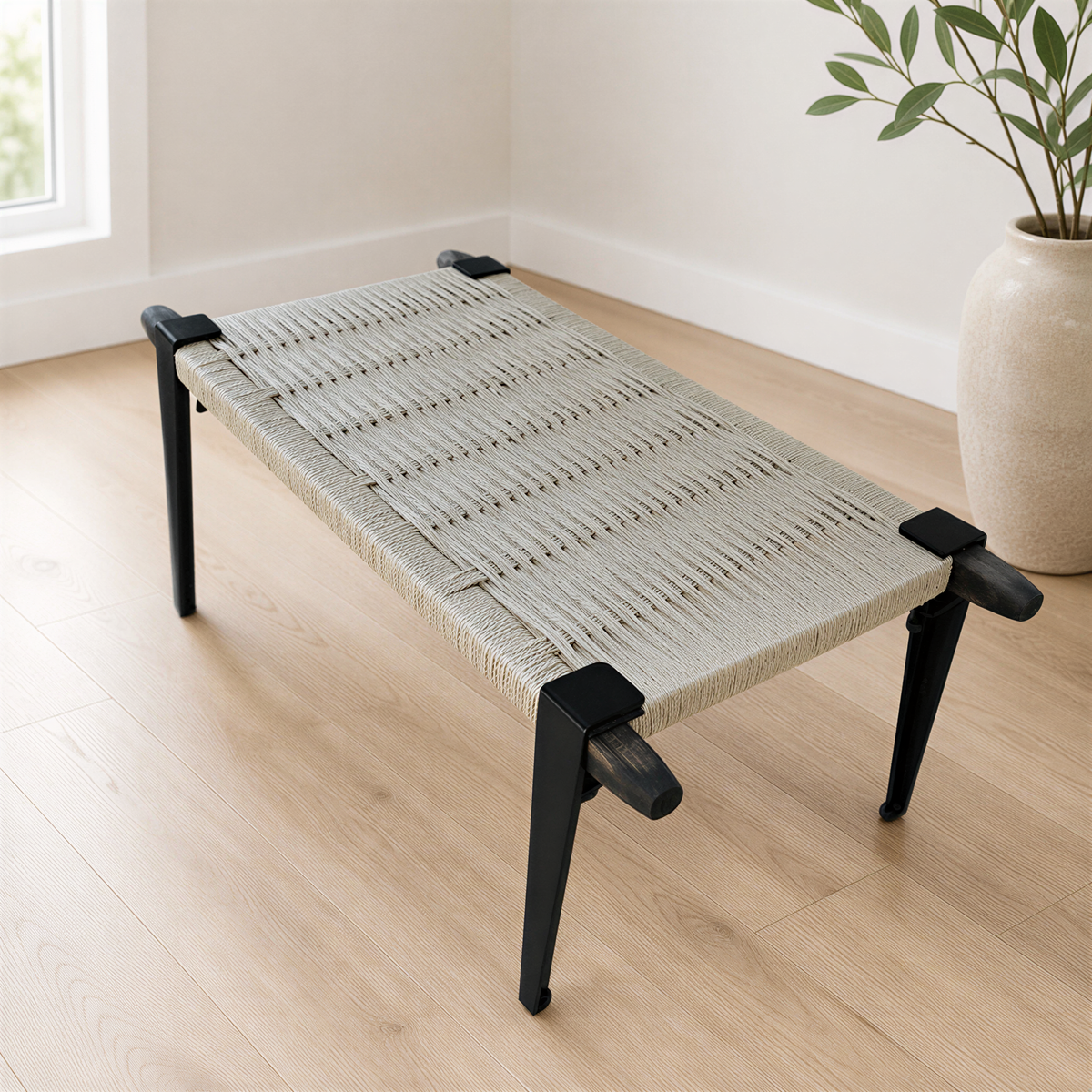 A woven bench with black metal legs sits on a wooden floor in a room with a window.