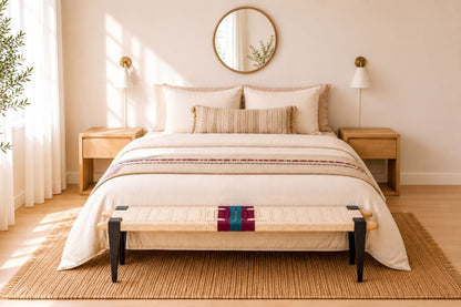 a serene and well-organized bedroom with a large bed, wooden nightstands, and a round mirror on the wall. The bed is dressed in a white comforter and has a colorful throw blanket on the bottom. The room has a warm, inviting atmosphere with a wooden floor and a rug.