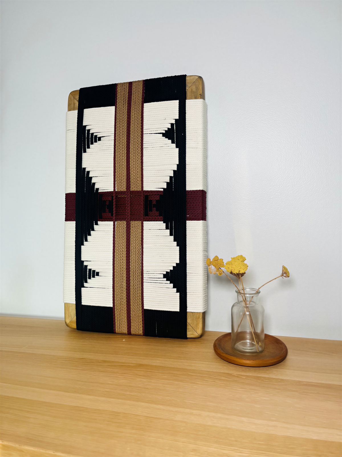 a wooden shelf with a decorative woven piece and a small vase with yellow flowers placed on it.