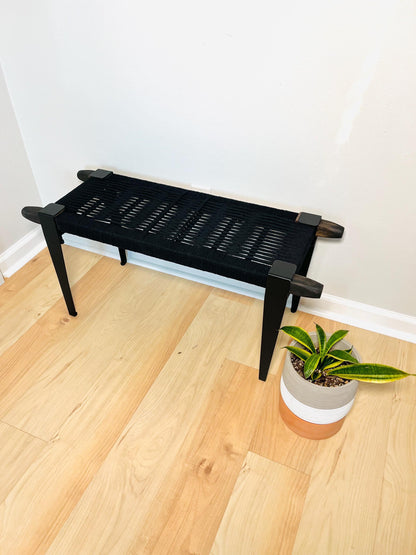 a black bench with a woven seat and legs, placed on a wooden floor next to a potted plant.