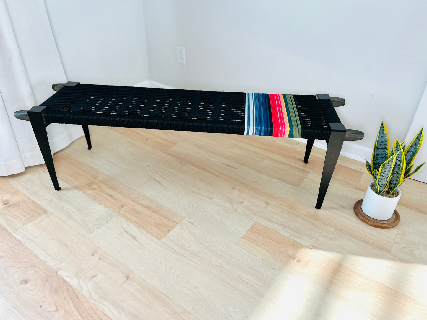 a black bench with a colorful striped cushion placed on a wooden floor, accompanied by a potted plant in the background.