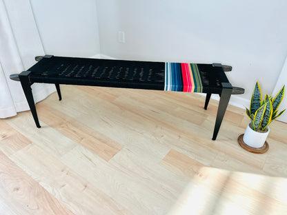 a black bench with a colorful striped cushion placed on a wooden floor, accompanied by a potted plant in the background.