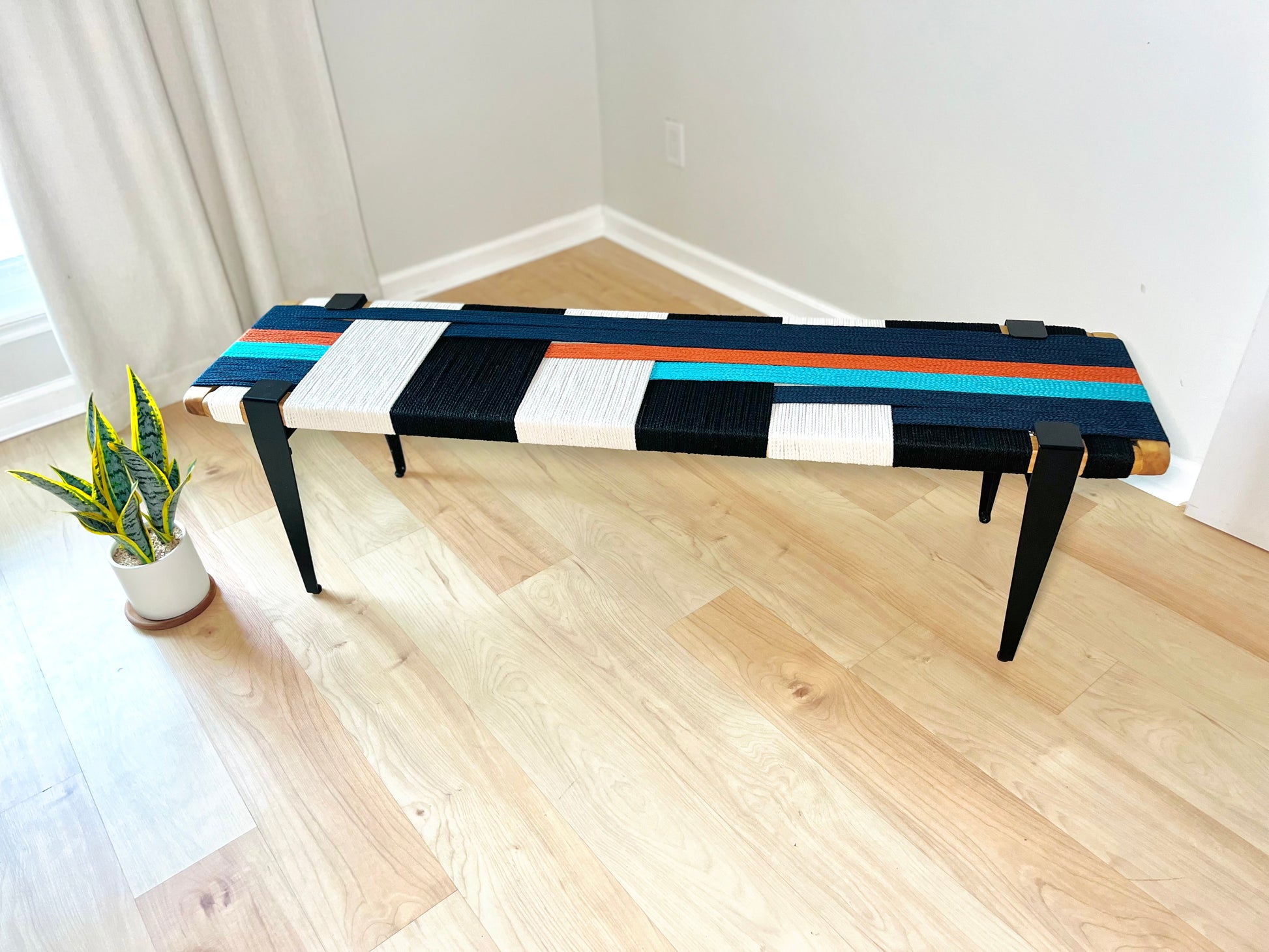 a modern, colorful bench with a geometric pattern of black, white, and orange stripes on the seat. The bench is placed on a wooden floor, and there is a potted plant nearby.
