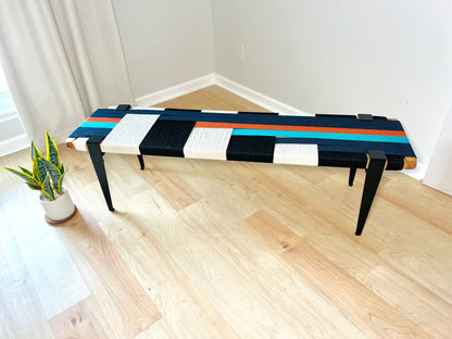 a modern, colorful bench with a geometric pattern of black, white, and orange stripes on the seat. The bench is placed on a wooden floor, and there is a potted plant nearby.