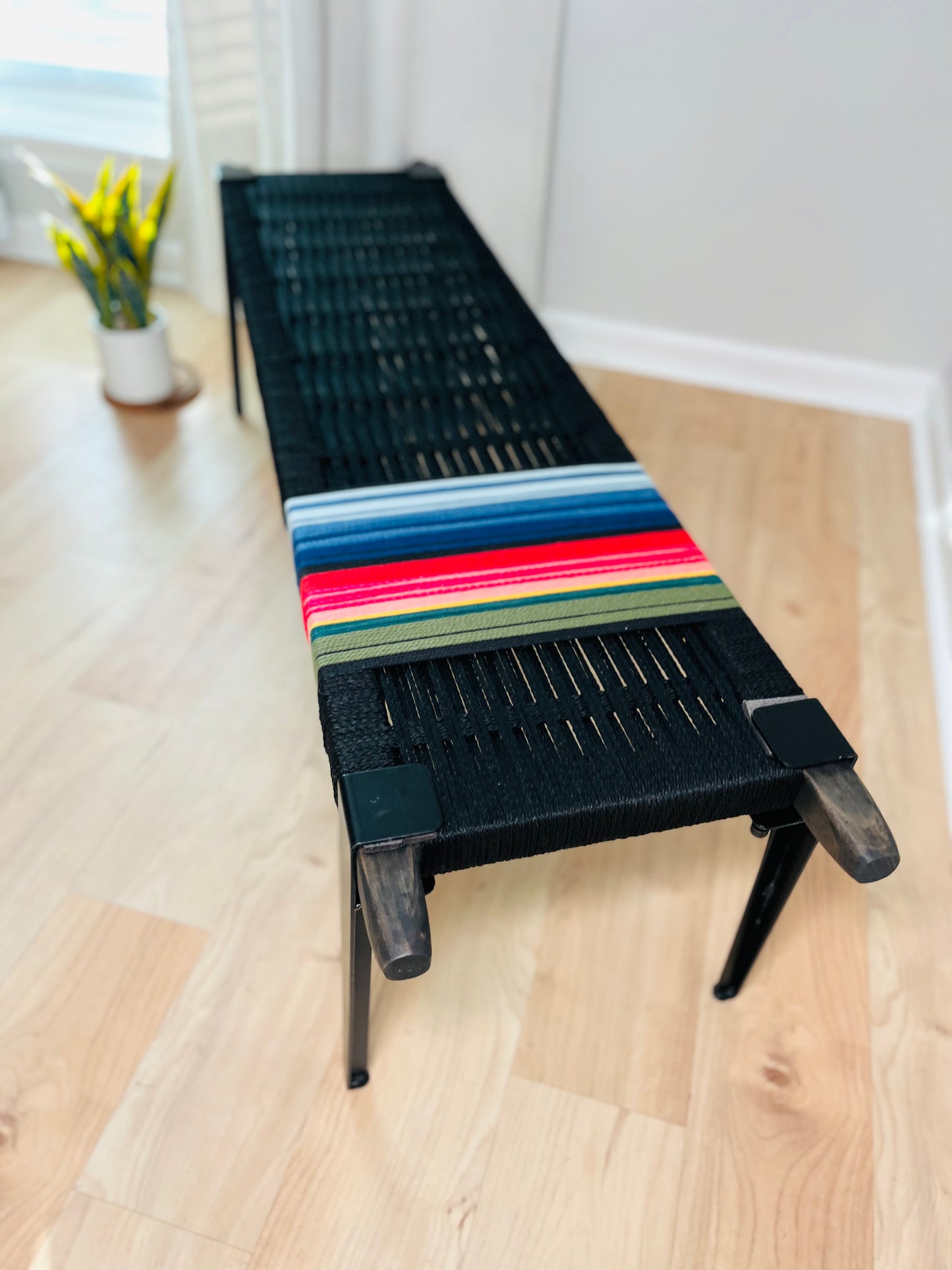 A black bench with a colorful woven blanket draped over it, placed on a wooden floor in a room with a window and a potted plant.