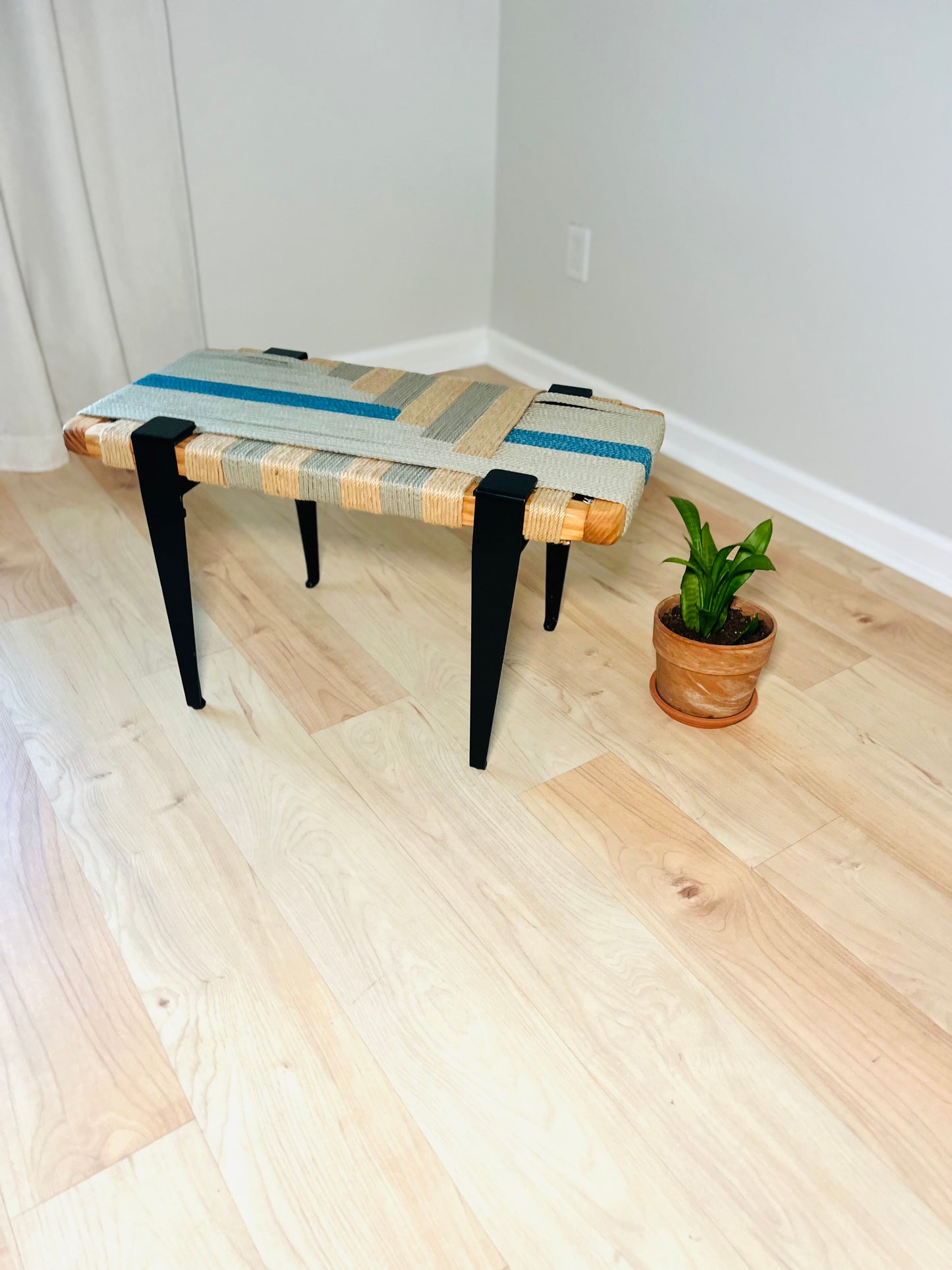 a woven bench with a striped cushion placed on a wooden floor, accompanied by a potted plant in the corner.