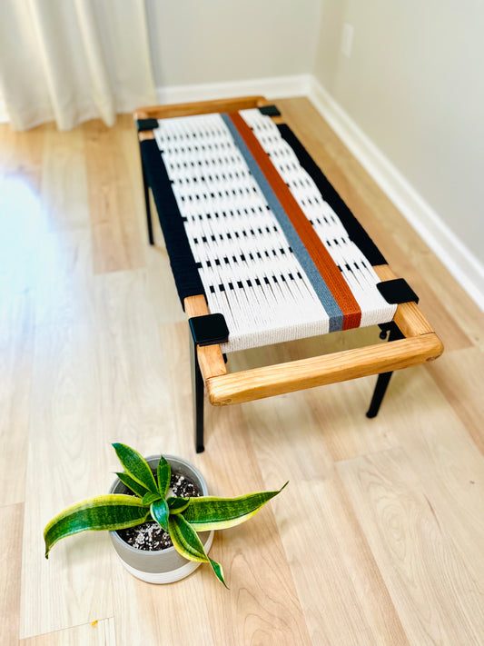 a woven bench with a wooden frame, placed on a wooden floor next to a potted plant.