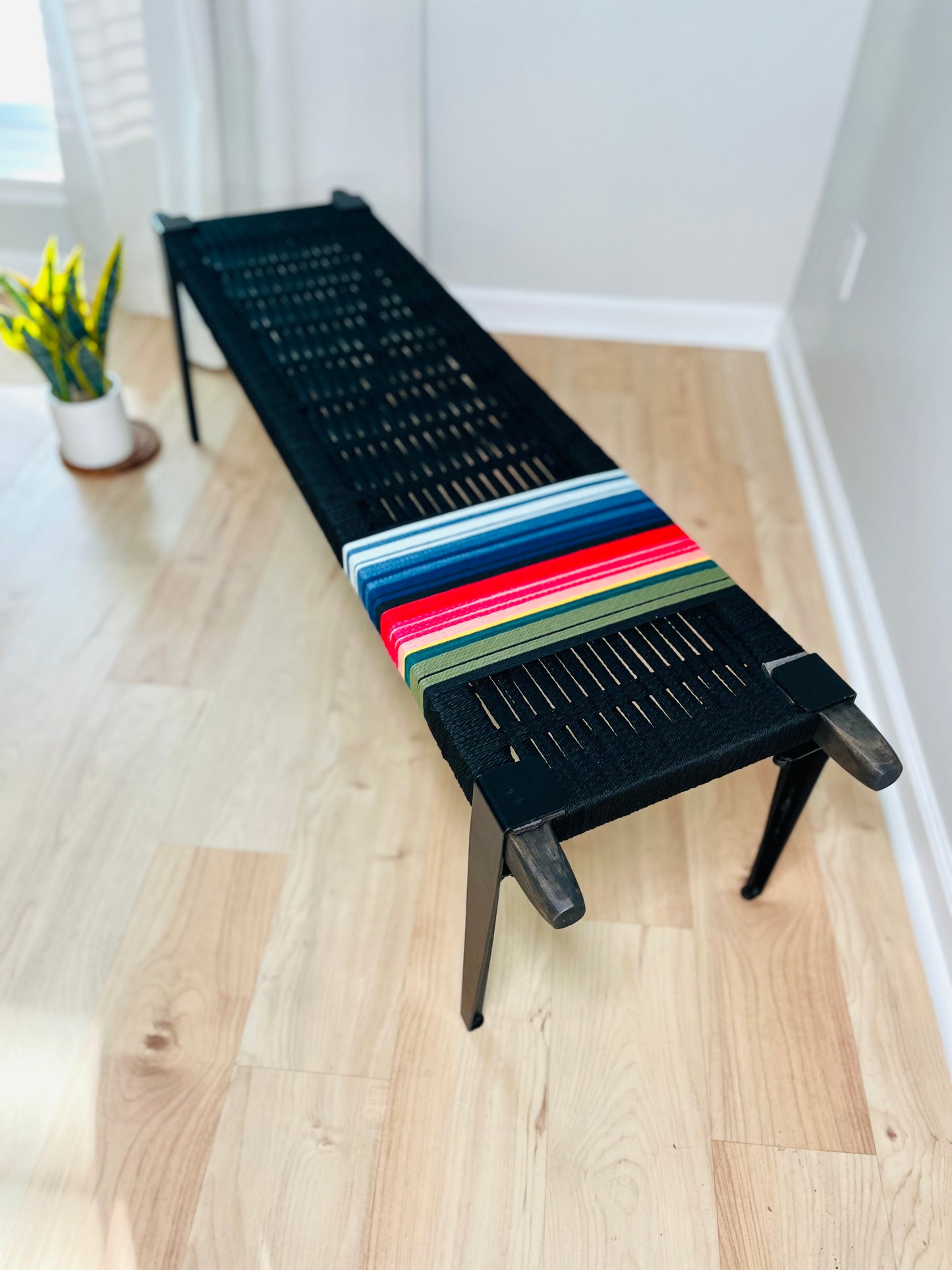 A black bench with colorful stripes on the seat is placed on a wooden floor in a room with a white wall and a window.
