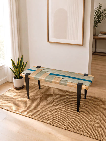a wooden bench with a woven seat and legs, placed on a woven rug in a room with a window and a plant.