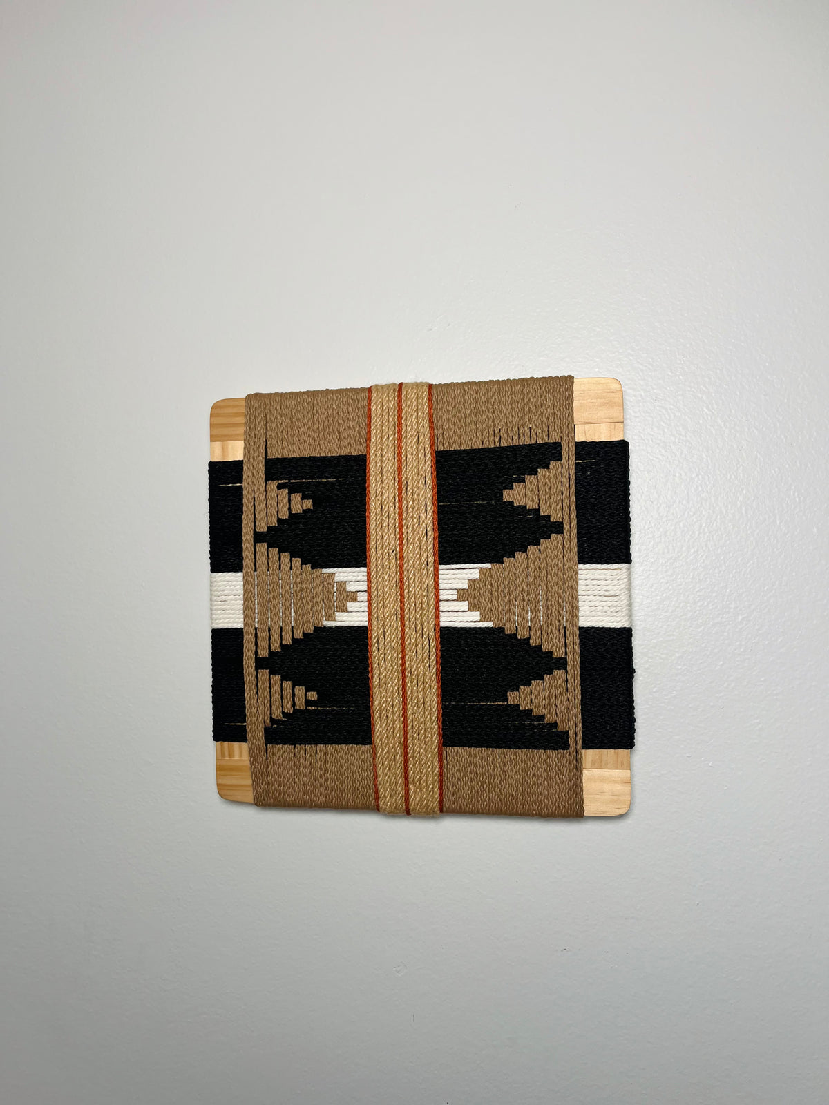 a woven piece of art, which appears to be a square-shaped textile or textile-like object. The artwork is displayed on a white wall, and it has a distinct geometric pattern with alternating black and white stripes.