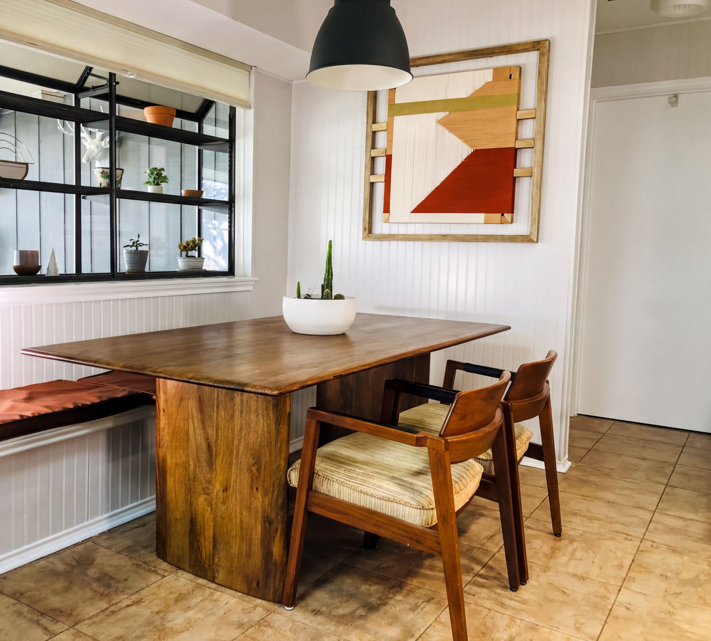 a cozy dining area with a wooden table and two matching chairs, one on each side. A large window with a black metal frame is visible, allowing natural light to fill the space. A potted plant is placed on the windowsill, adding a touch of greenery to the room. A framed abstract artwork hangs on the wall above the table, complementing the overall decor.