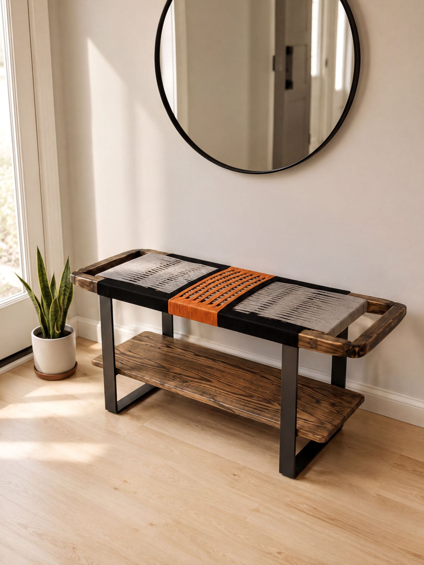 a modern, minimalist design with a wooden bench and a round mirror on the wall. The bench has a black and orange woven seat, and the mirror is mounted above it. The room has a clean, white color scheme with a wooden floor, and a potted plant is visible in the corner.