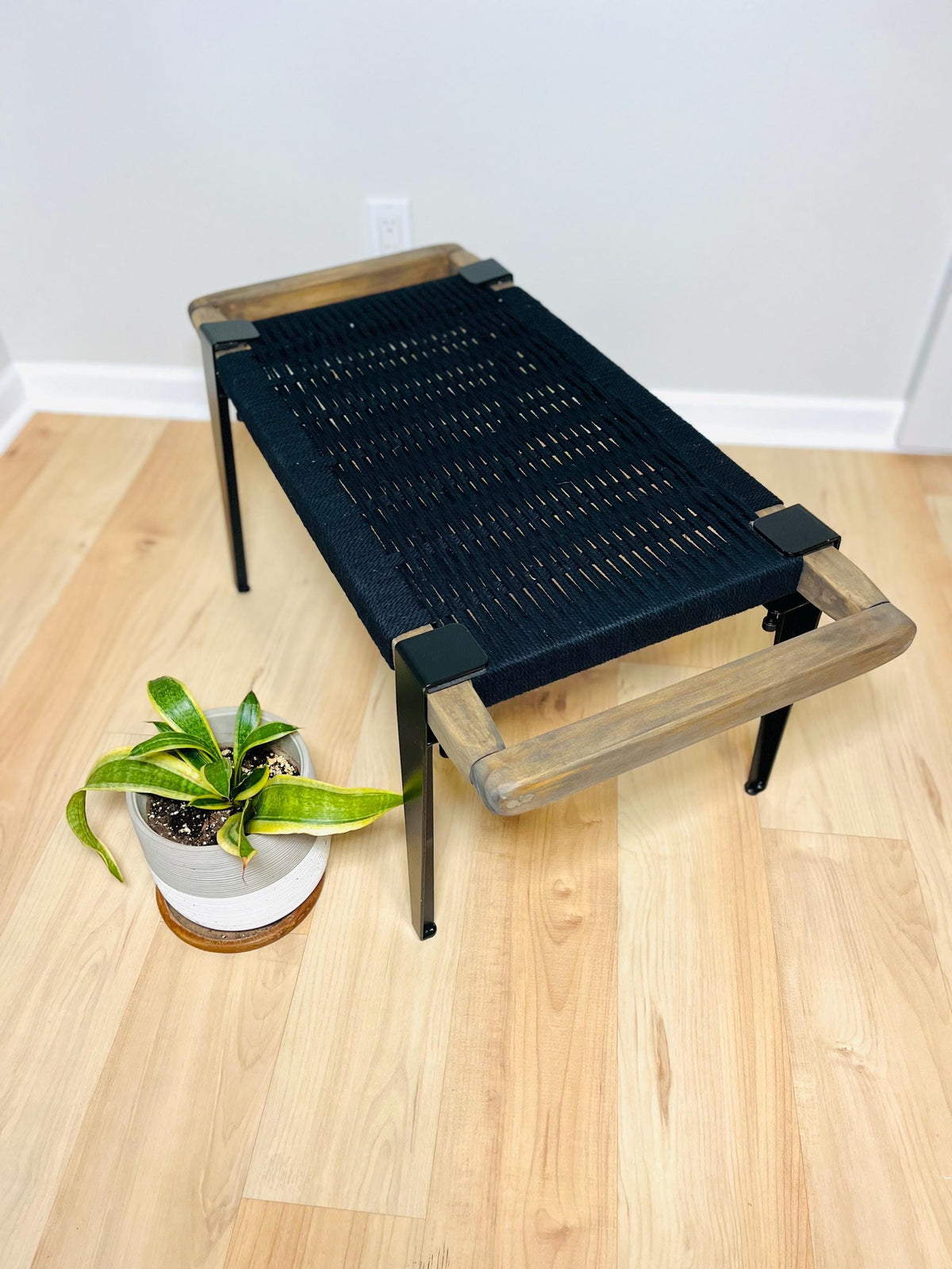 a black woven bench with a wooden frame, placed on a wooden floor next to a potted plant.