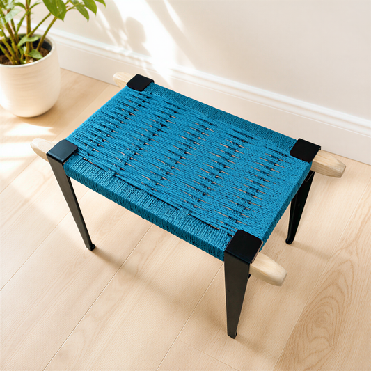 A blue woven bench with black metal legs sits on a wooden floor, with a potted plant visible in the background.