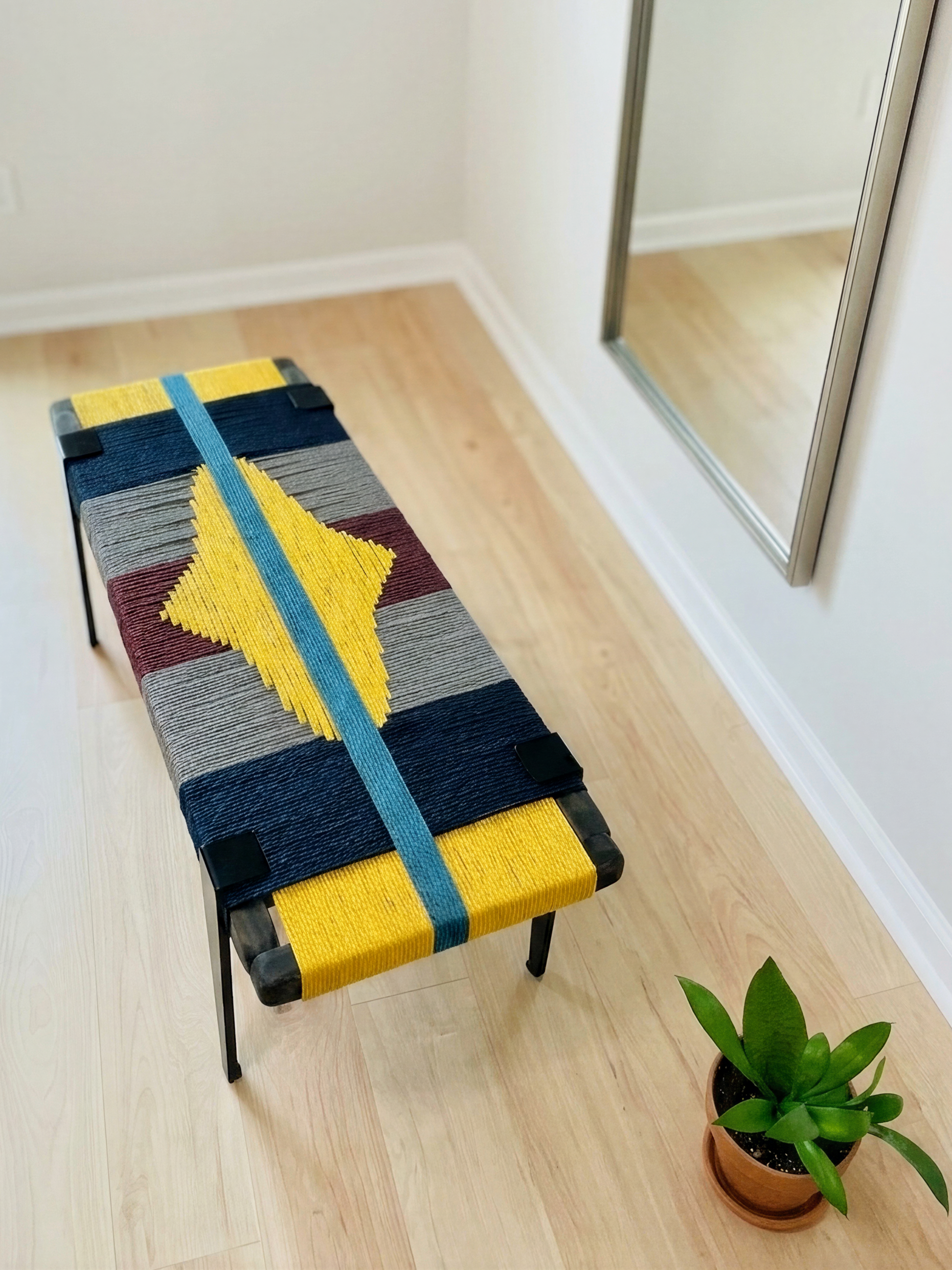 a colorful bench with a yellow and blue design, placed on a wooden floor in a room with a mirror and framed pictures on the wall.