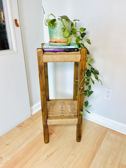 A small wooden side table with a woven seat and a potted plant on top, placed on a wooden floor against a white wall.